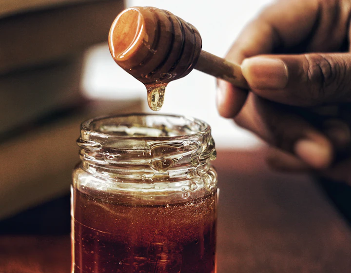 Tangan mengangkat sendok madu kayu yang meneteskan madu ke botol kaca terbuka
