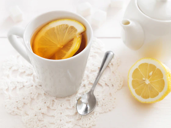 Cangkir putih berisi teh dengan irisan lemon, sendok, dan setengah lemon di sampingnya